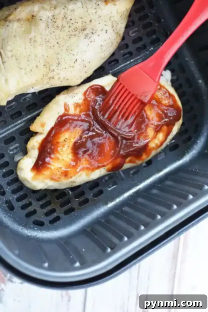 Perfect Air Fryer BBQ Chicken Breast 7 A piece of air fryer BBQ chicken is being brushed with barbecue sauce using a red silicone brush inside the basket. Another piece of seasoned chicken sits tantalizingly in the background, ready to be savored.