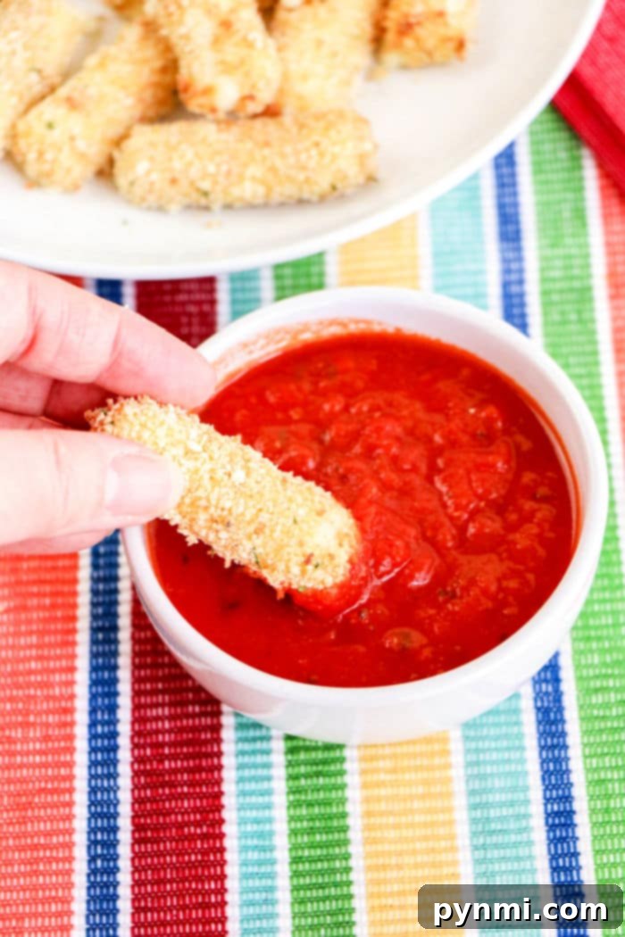 Crispy Homemade Air Fryer Mozzarella Sticks 8 A hand dipping a crispy homemade mozzarella stick into a bowl of vibrant red marinara sauce, with more sticks in the background.