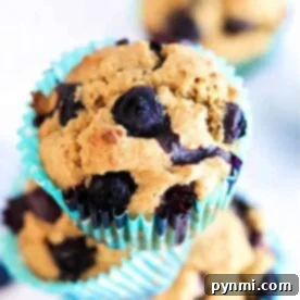 A blueberry bran muffin with a golden crust sits in a turquoise wrapper against a blurred background.