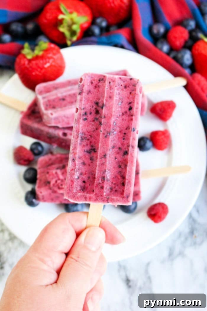 A hand holds a vibrant berry popsicle, surrounded by other popsicles and fresh berries on a plate, highlighting the delightful and easy process of how to make homemade popsicles using frozen fruit.