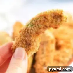 Close-up of a hand holding an air fryer avocado fry. The avocado is coated in a crispy, golden-brown crust, with more pieces in soft focus in the background.