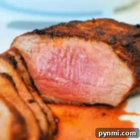 Close-up of a cooked air fryer tri-tip steak, sliced to reveal a pink, juicy interior. The steak boasts a seasoned, grilled crust with some juices pooled around it on the white plate.