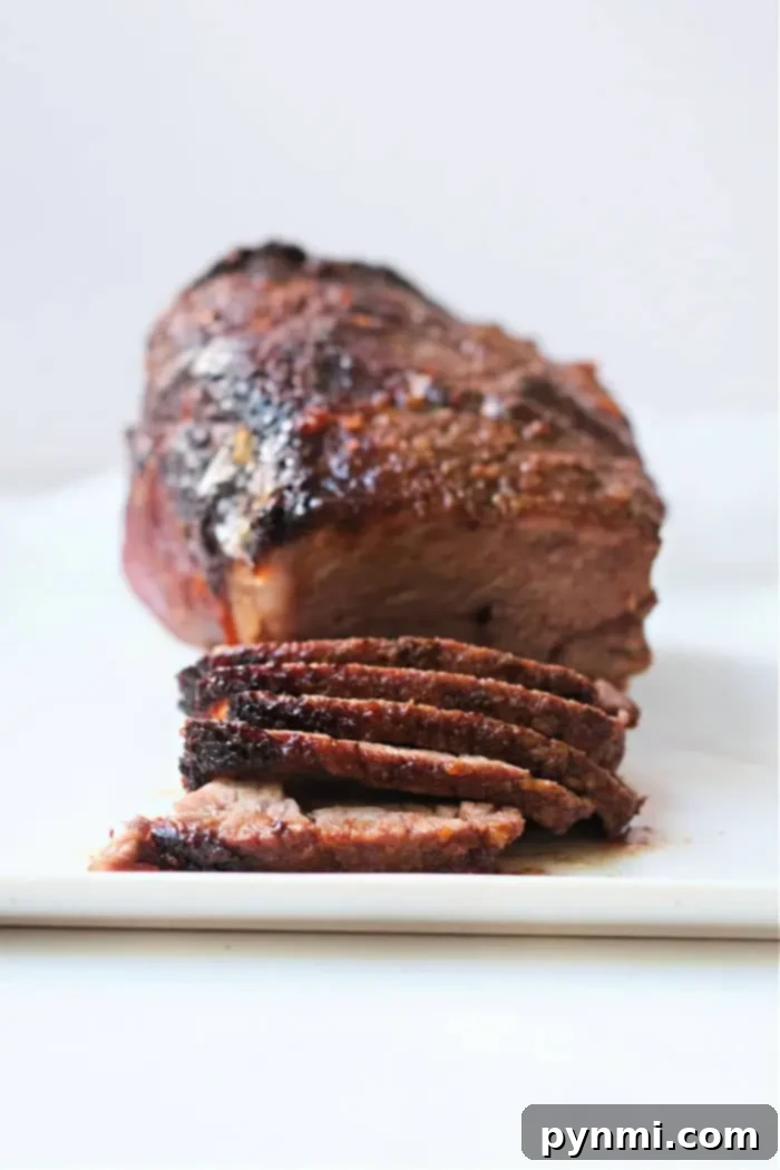 Image of a roasted beef brisket with a dark, crispy crust, sitting on a white cutting board. The brisket is partially sliced, showing its interior texture and juiciness. Prepared using the air fryer roast beef method, the background remains neutral and plain.