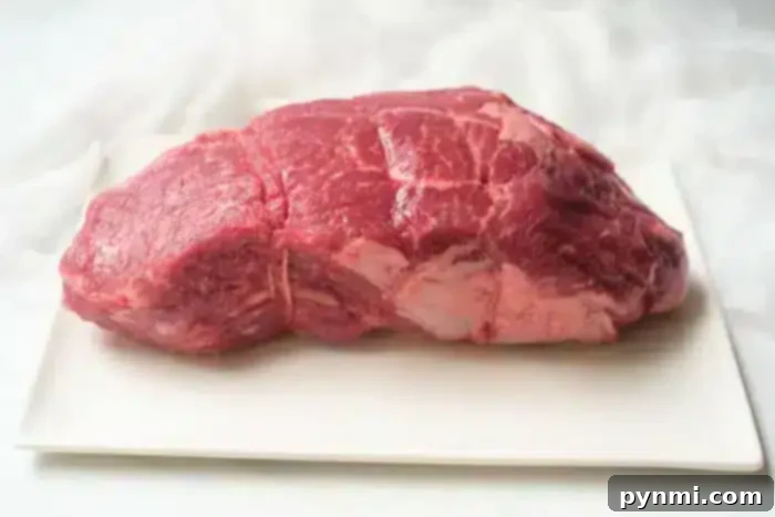 A raw cut of beef placed on a rectangular white ceramic platter, showcasing its visible marbling and fat. The background is light and softly blurred, highlighting the red color and texture of what promises to be a delectable air fryer roast beef.