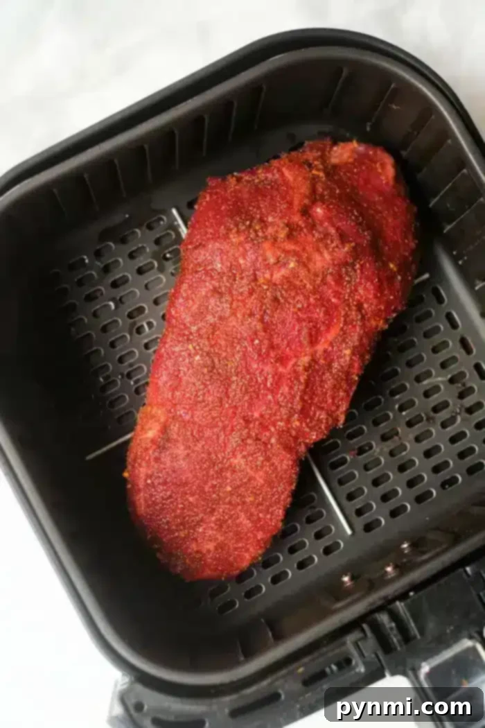 A piece of seasoned roast beef is placed in an air fryer basket. The meat has a reddish-brown crust, suggesting it has been coated with a spice rub. The air fryer basket has a dark non-stick surface with perforations for optimal air circulation.
