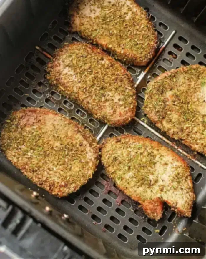 Perfect Air Fryer Pork Steak 2 Five seasoned pork steaks are evenly spaced in an air fryer basket. They are covered in a mixture of herbs and spices, giving them a crispy, golden-brown appearance.