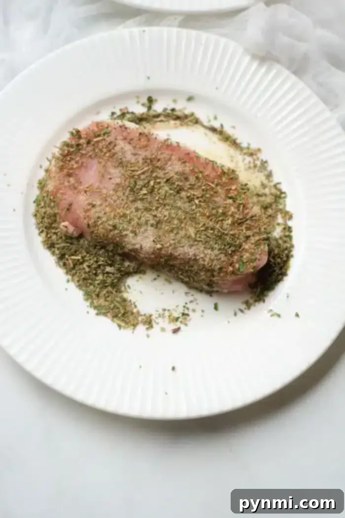 Perfect Air Fryer Pork Steak 4 A pork steak, seasoned with a variety of herbs, rests on a white, ridged plate. The finely chopped herbs blanket the top of the meat and spill over onto the plate. Perfect for an air fryer, the background sets a light, textured scene.