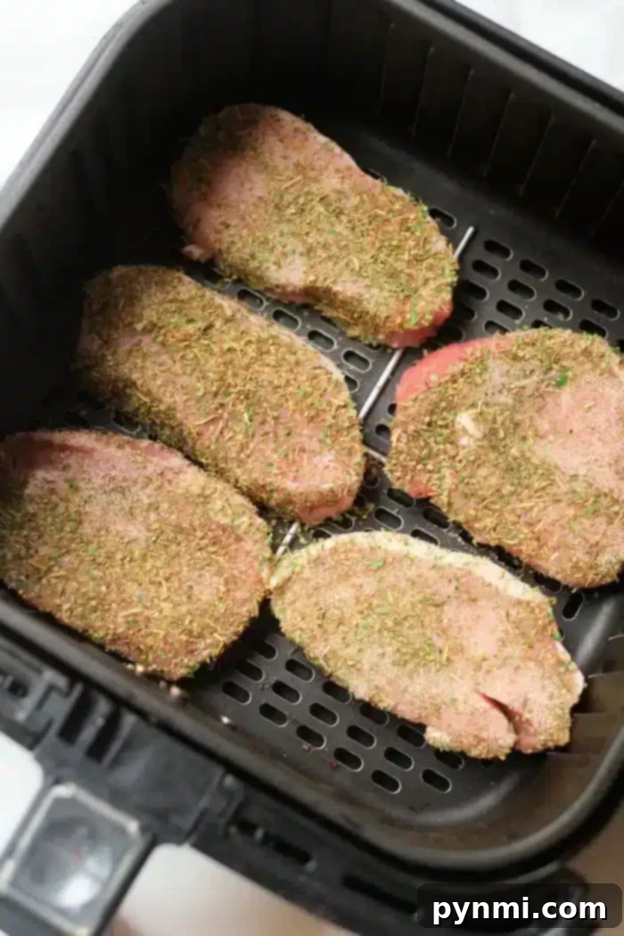 Perfect Air Fryer Pork Steak 5 Five uncooked, seasoned pork steaks are placed in an air fryer basket, ready for cooking. The meat is covered with a mixture of herbs and spices.