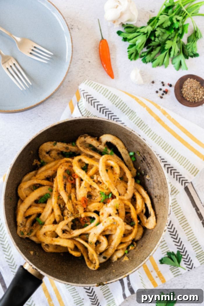 A pan of fried calamari with herbs and spices rests on a striped cloth beside forks, garlic, pepper, and parsley, much like an inviting setting for learning how to cook calamari.