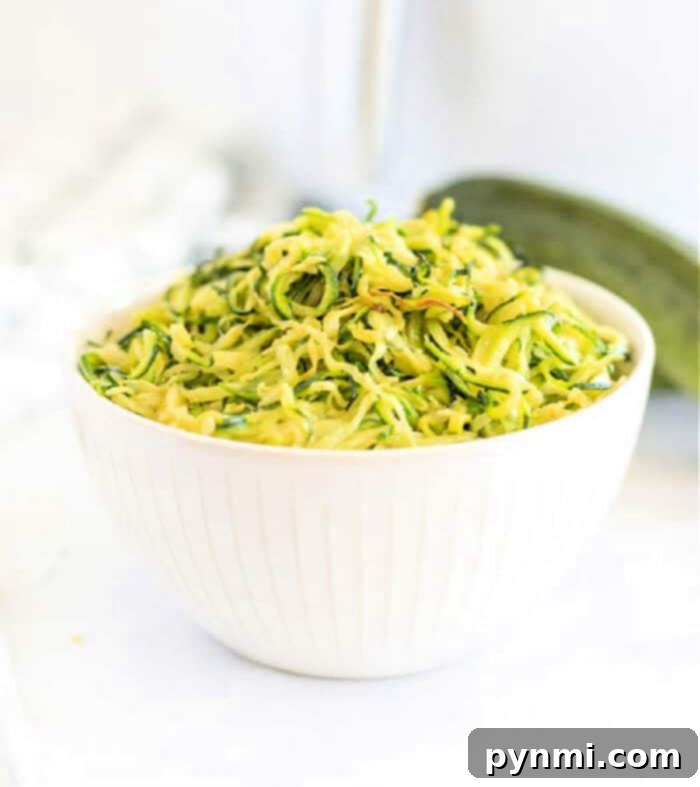 White bowl filled with shredded zucchini on a white surface, perfect for making air fryer zucchini noodles, with a fresh zucchini in the background.