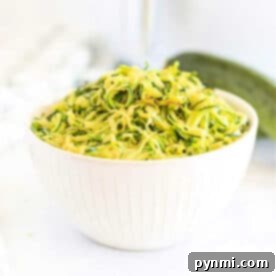 White bowl filled with shredded zucchini on a white surface, perfect for making air fryer zucchini noodles, with a fresh zucchini in the background.
