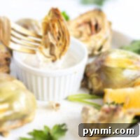 A fork holds an air fryer artichoke above a dipping sauce on a white plate, garnished with lemon and parsley.
