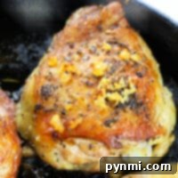 Close-up of a seasoned, golden-brown garlic butter chicken thigh in a skillet, topped with minced garlic and herbs.
