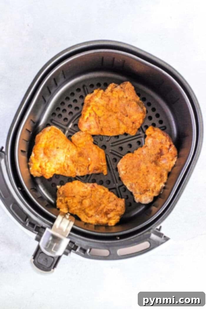 Four perfectly seasoned chicken thighs are nestled in an air fryer basket, their crispy golden edges promising a delightful and healthier alternative to traditional fried chicken. A testament to easy air fryer recipes.