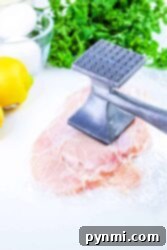 Raw chicken breast being tenderized with a metal mallet, surrounded by eggs, parsley, and a lemon.