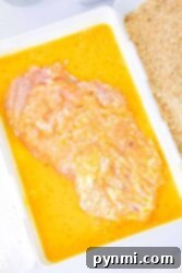 Raw chicken breast marinating in a yellow mixture, next to breadcrumbs in a white tray.