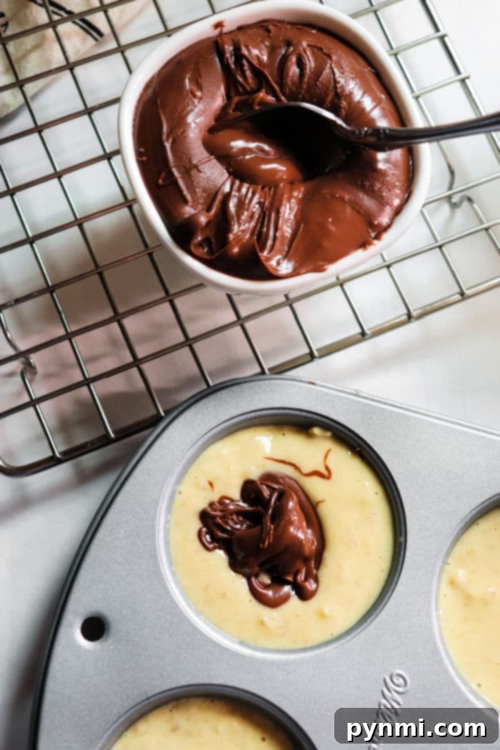 Nutella Swirl Banana Muffins 5 Muffin batter in a baking tin with a luscious dollop of chocolate Nutella on top, positioned next to a bowl of melted chocolate on a cooling rack. This image beautifully captures the preparation of delectable banana Nutella muffins, hinting at the indulgent treat to come.