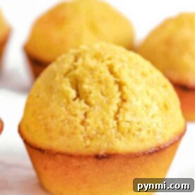 Golden Corn Muffins 5 Close-up of a golden-brown cornmeal muffin with a slightly cracked top, sitting on a white surface.