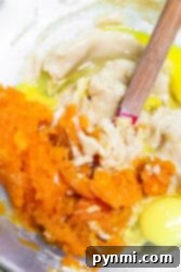 Mixing bowl containing raw ingredients: mashed orange sweet potato, an egg, and dough being combined with a wooden spoon for a delightful sweet potato muffin recipe.