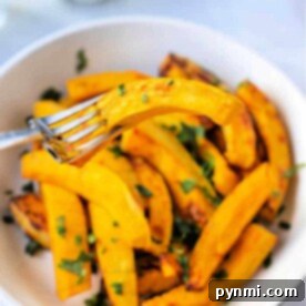A bowl of roasted pumpkin wedges, perfectly crisped in the air fryer, is garnished with herbs. A fork holds one golden wedge above the bowl.