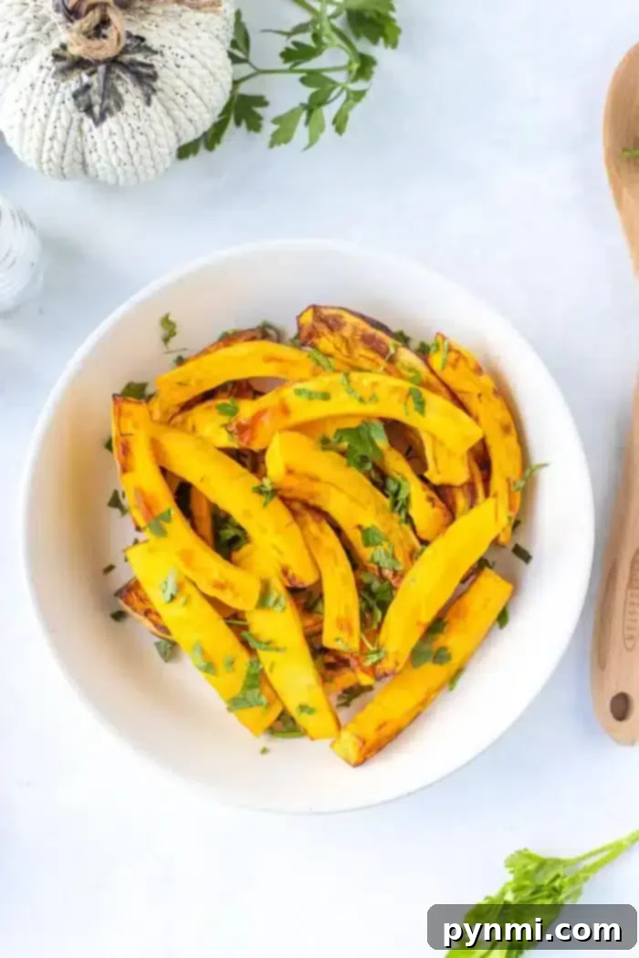 Air Fryer Roasted Pumpkin 8 A white bowl contains roasted yellow squash garnished with chopped green herbs on a white surface. There is a wooden spoon on the right, and part of a decorative pumpkin and parsley can be seen in the top left corner, hinting at air fryer pumpkin slices to complete the autumnal scene.
