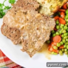 A quick meatloaf recipe served elegantly: a plate with sliced meatloaf, topped with rich gravy, accompanied by vibrant peas and carrots, and a slice of bread on a pristine white dish.