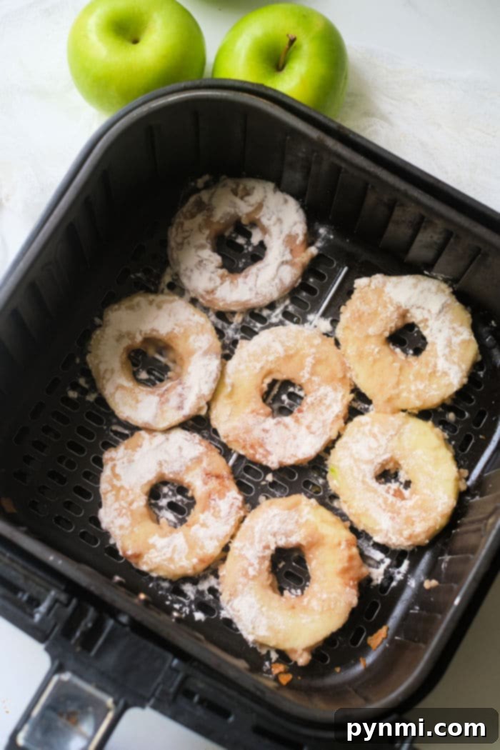 Air fryer apple rings, perfectly crisp and golden, nestle in the basket while two fresh green apples stand in the background, hinting at their fresh origins.