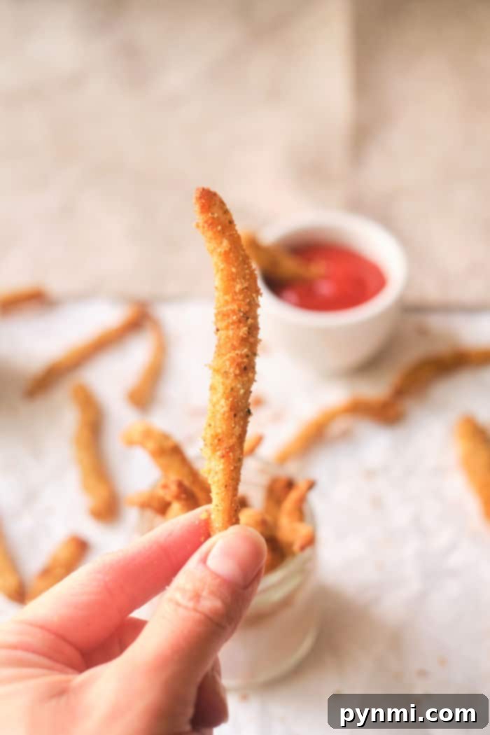 Close-up of a hand holding a crispy chicken fry, with more fries and dipping sauce in the background, reminiscent of chicken fries cooked to perfection in an air fryer.