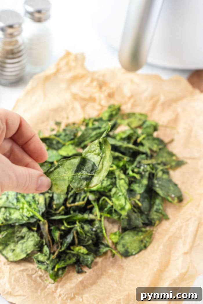A hand holds a crisp spinach leaf over a pile of perfectly air-fried spinach on parchment paper, showcasing the delicious result of the air fryer spinach method.