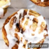 Close-up of a cinnamon roll muffin topped with white icing. The golden brown treat reveals its swirled layers, dusted with sprinkled cinnamon, as the icing drips down the sides. Two partial muffins are visible in the background on a marble surface.