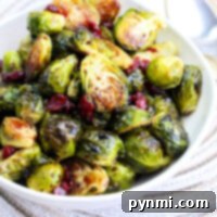 A white bowl filled with roasted Brussels sprouts and cranberries drizzled in a rich balsamic glaze rests on a woven placemat.