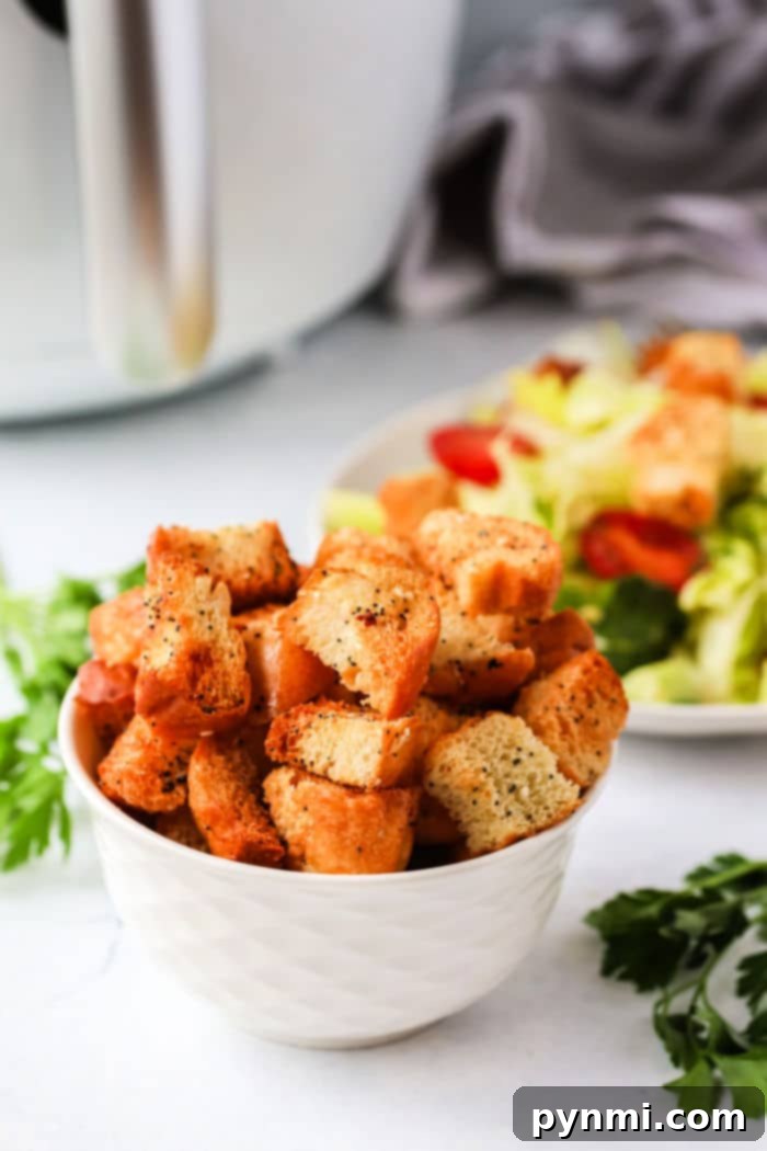 A bowl of perfectly seasoned air fryer croutons sits beside a fresh salad with tomatoes and lettuce, on a white countertop with scattered herbs.