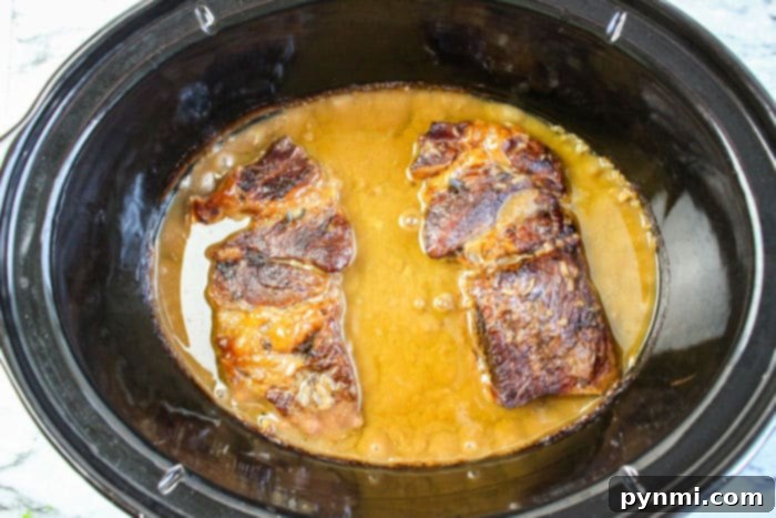 Two browned chuck roast pieces and gravy simmering in a black slow cooker, creating rich flavors
