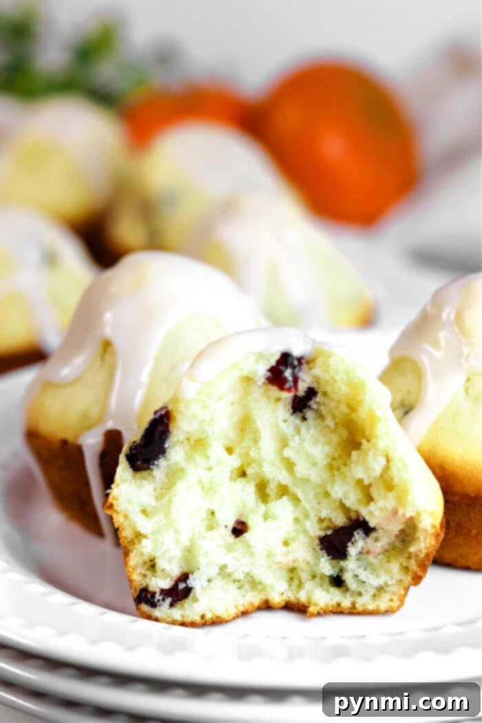 A sliced, healthy cranberry muffin with yogurt and icing sits on a plate, while more muffins and fresh oranges create an inviting background, highlighting the vibrant colors and appealing texture of the muffins.