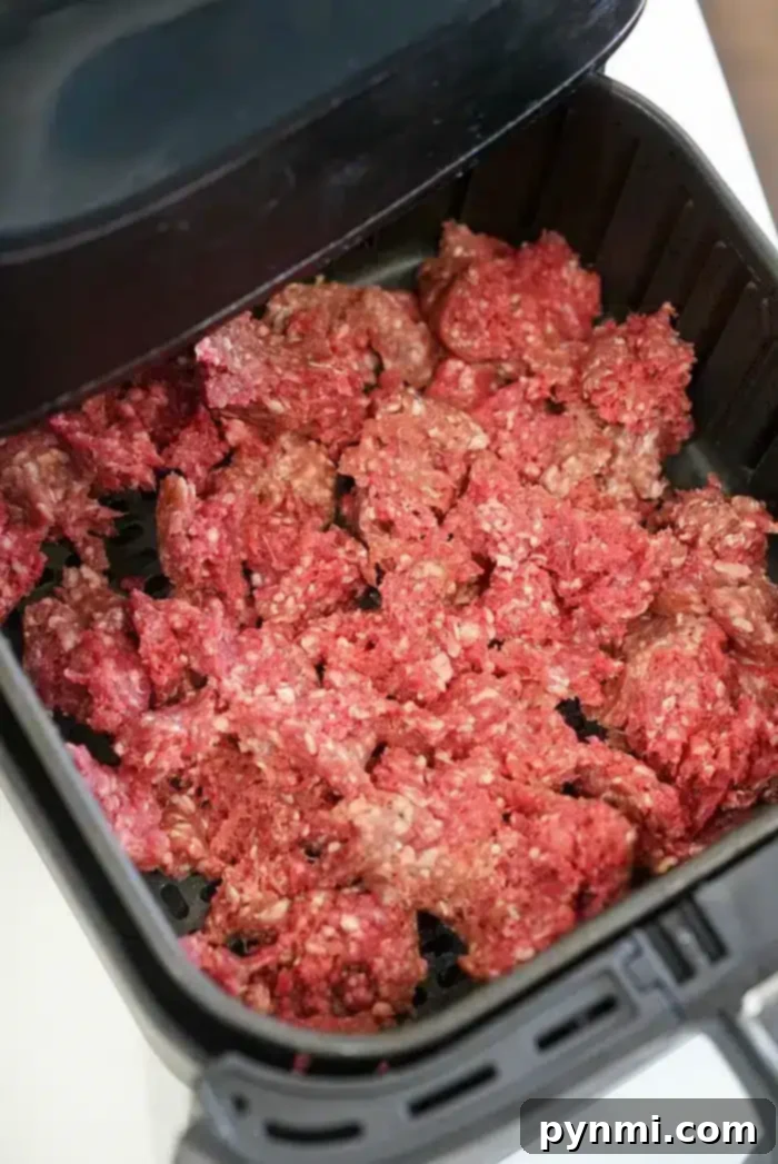 Ground beef is evenly spread out in an air fryer basket, perfectly prepared for cooking. The air fryer lid is partially visible in the upper portion of the image, showcasing an ideal setup for preparing various air fryer beef recipes.