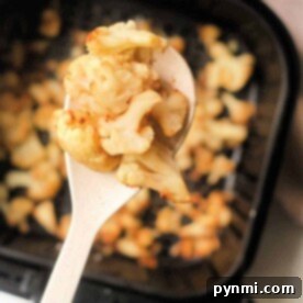 Close-up of roasted cauliflower florets on a spoon, showcasing the perfect transformation from frozen to crispy in an air fryer basket filled with more golden goodness.