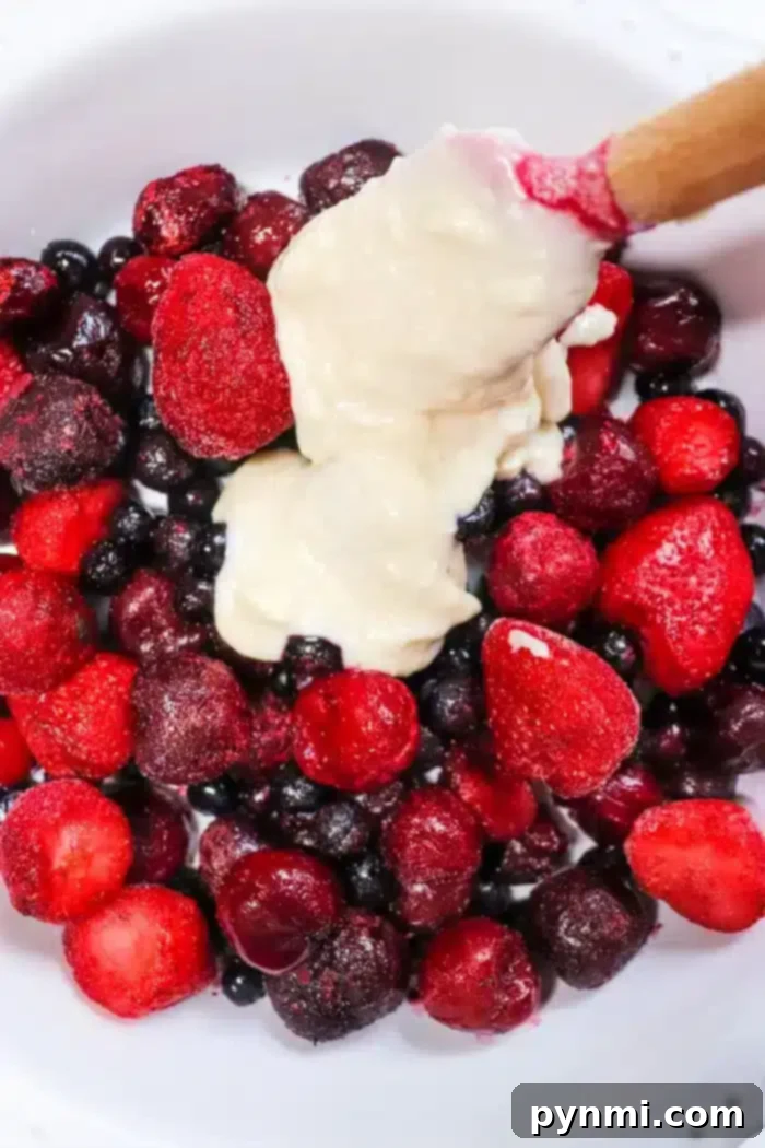 Effortless 5-Ingredient Bisquick Blackberry Cobbler 5 A bowl of assorted berries, including strawberries, blueberries, and blackberries, topped with a dollop of creamy batter reminiscent of a Bisquick cobbler. A wooden spoon is partially dipped into the mixture.