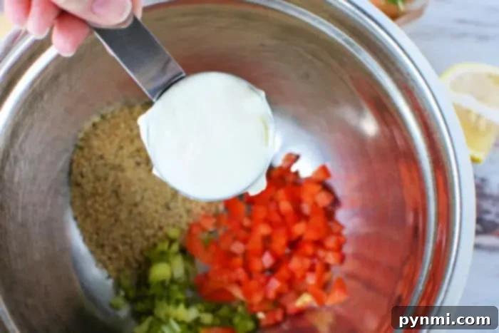 Crispy Air-Fried Crab Cakes 3 A hand is holding a measuring cup filled with mayonnaise over a mixing bowl. Inside the bowl are ingredients for Air Fryer Crab Cakes, including breadcrumbs, chopped red bell pepper, and chopped celery.