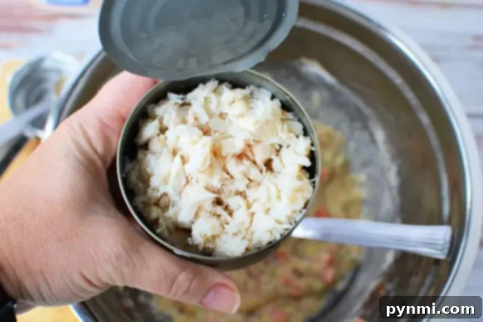 A hand is holding an open can of crab meat over a stainless steel mixing bowl. A spoon is visible inside the bowl, which contains a mixture of chopped vegetables and other ingredients for Air Fryer Crab Cakes.