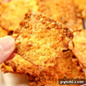 A close-up of a hand holding a square, crispy cheese cracker with jagged edges. Several other crackers are visible in the background on a plate, resembling delightful pasta chips with their golden-brown hue and textured surface.
