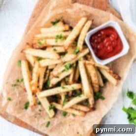 Crispy air fryer jicama fries garnished with herbs, served on parchment with a small dish of ketchup on a wooden board.
