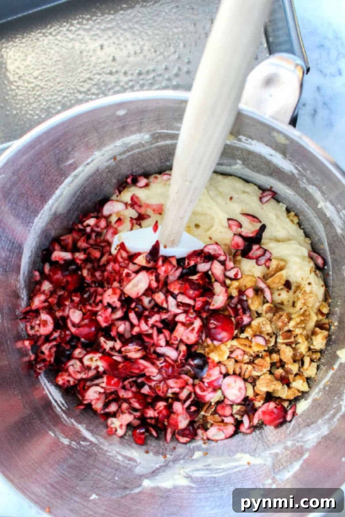 Chopped cranberries and walnuts are being mixed into batter for delicious banana cranberry bread in a metal bowl with a spatula.