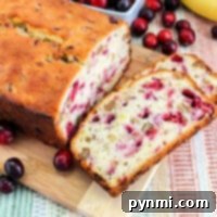 Sliced banana cranberry bread with walnuts rests on a wooden board, surrounded by fresh cranberries and a perfectly ripe banana, ready to be enjoyed.