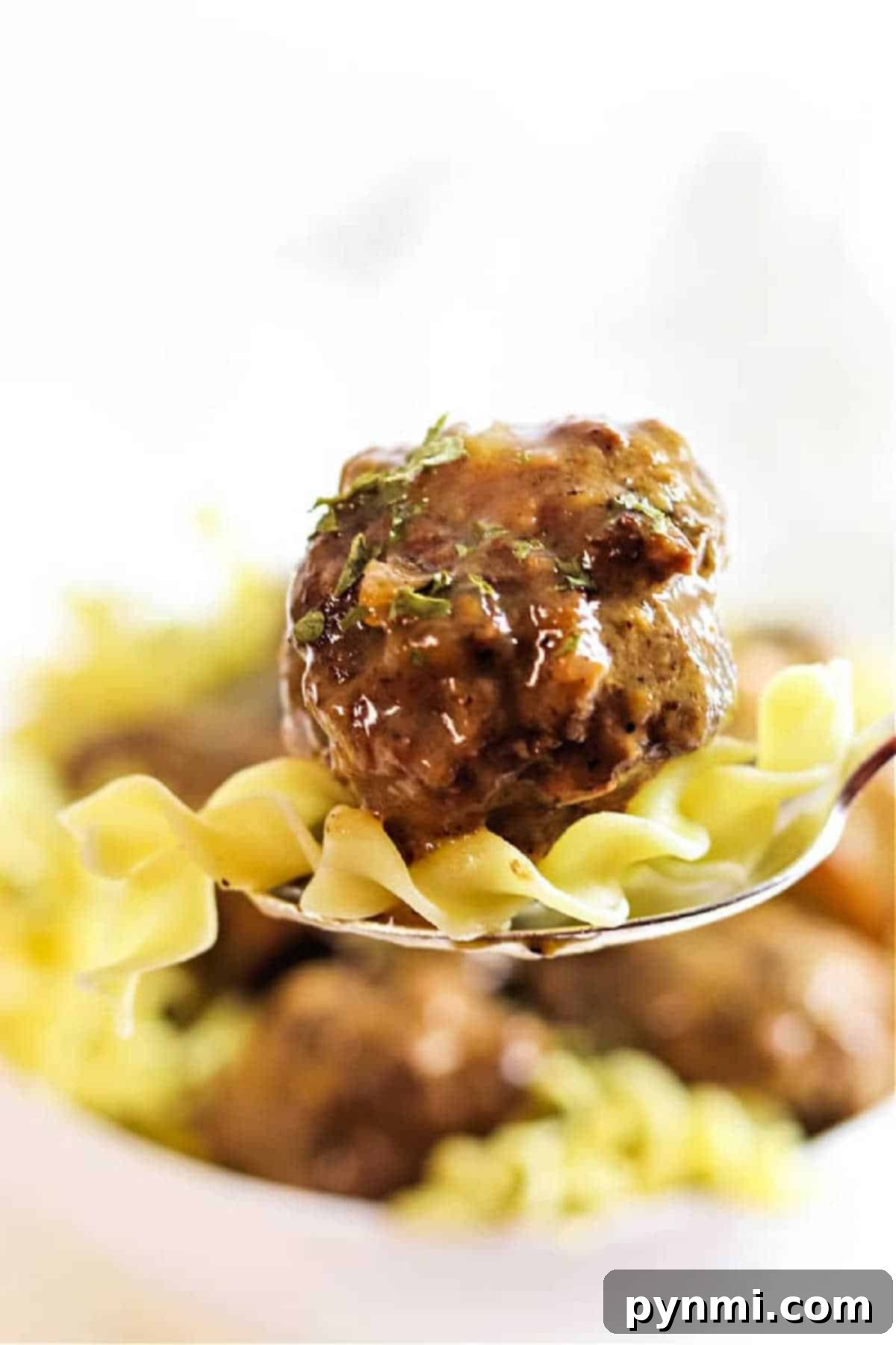 Close-up of a fork with a meatball and noodles, garnished with herbs, evokes the comforting flavors of Crockpot Meatballs and Gravy.