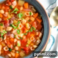 A bowl of healthy minestrone soup with pasta, vegetables, and herbs, simmered to perfection in a crockpot, sits next to a gold spoon on a patterned napkin.