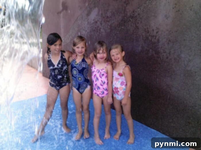 Four young children in swimsuits stand and smile under a water feature at the Lake Skinner splash pad. A water stream is visible on the left, with the kids standing on a blue surface against a textured wall, highlighting the joy of the experience.
