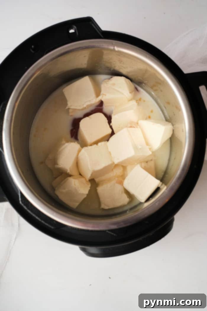 Large cubes of softened cream cheese partially melted in a metal pressure cooker pot, surrounded by a vibrant, creamy buffalo chicken dip liquid, hinting at the rich and smooth texture to come.