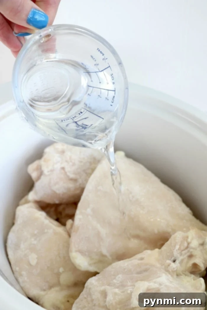 Mastering Tender Frozen Meat 13 A hand holding a measuring cup pours water over frozen chicken breasts in a white crockpot, illustrating the beginning of a frozen chicken slow cooker recipe.