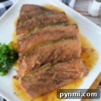 Sliced roast beef on a white rectangular plate, garnished with fresh parsley. A checkered black and white napkin and a salt shaker are placed nearby on a wooden surface. Juices from the frozen roast in the oven are visible on the plate.