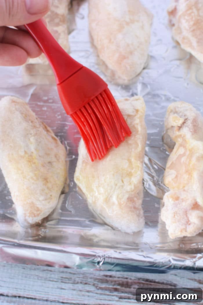 A hand applies oil to frozen chicken wings on a foil-lined baking sheet.